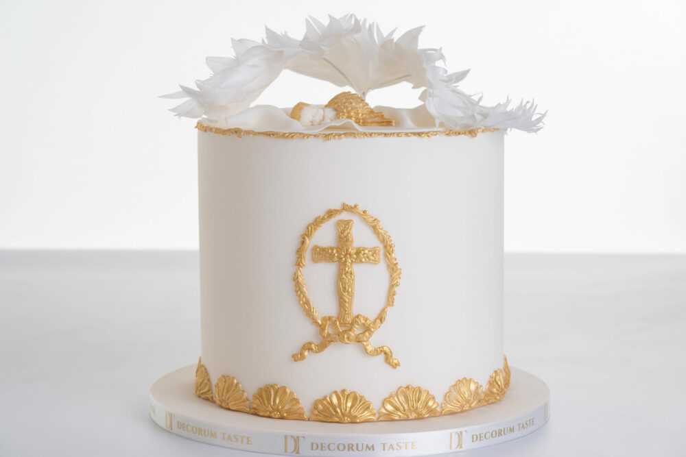 Single tier luxury contemporary, customisable, celebration cake, Christening Cake by Decorum Tastes in ivory with edible gold decorative detail, a hand-crafted baby in sugar paste sleeps underneath angel wings made from rice paper. Embellished with a golden cross with floral detail, gold bow and decorative border.