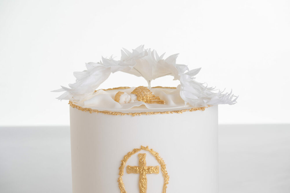 Top of luxury contemporary, customisable, celebration cake, Christening Cake by Decorum Tastes in ivory with edible gold decorative detail, a hand-crafted baby in sugar paste sleeps underneath angel wings made from rice paper.