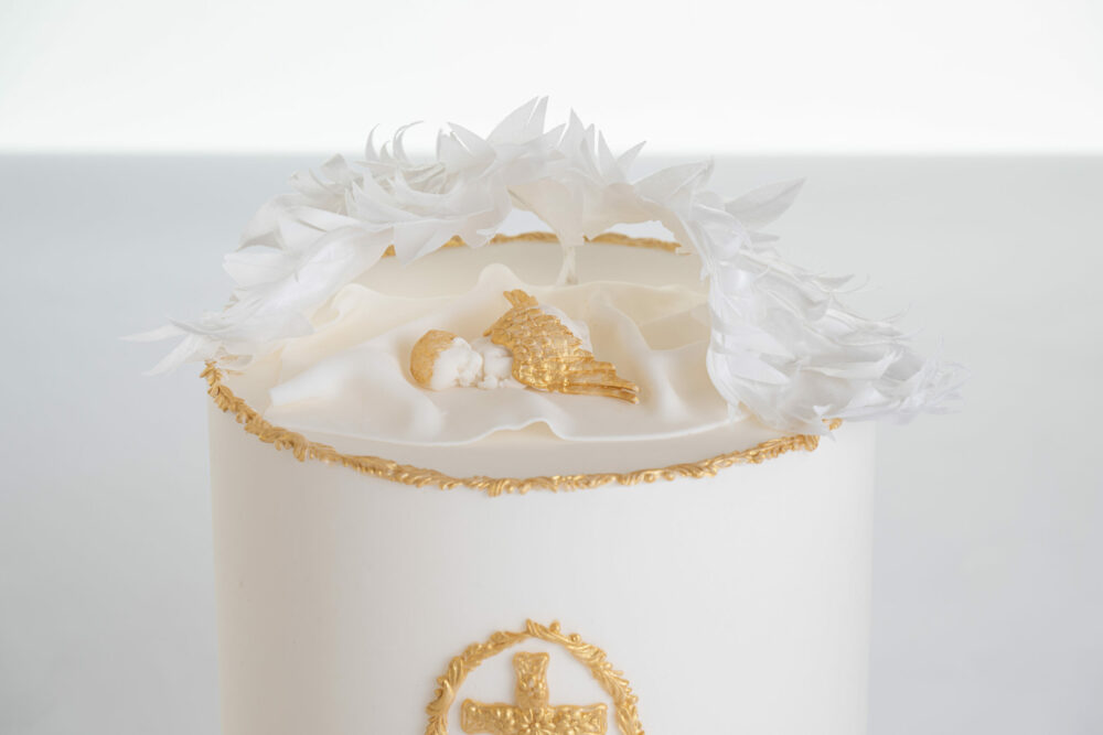 Top of luxury contemporary, customisable, celebration cake, Christening Cake by Decorum Tastes in ivory with edible gold decorative detail, a hand-crafted baby in sugar paste sleeps underneath angel wings made from rice paper.