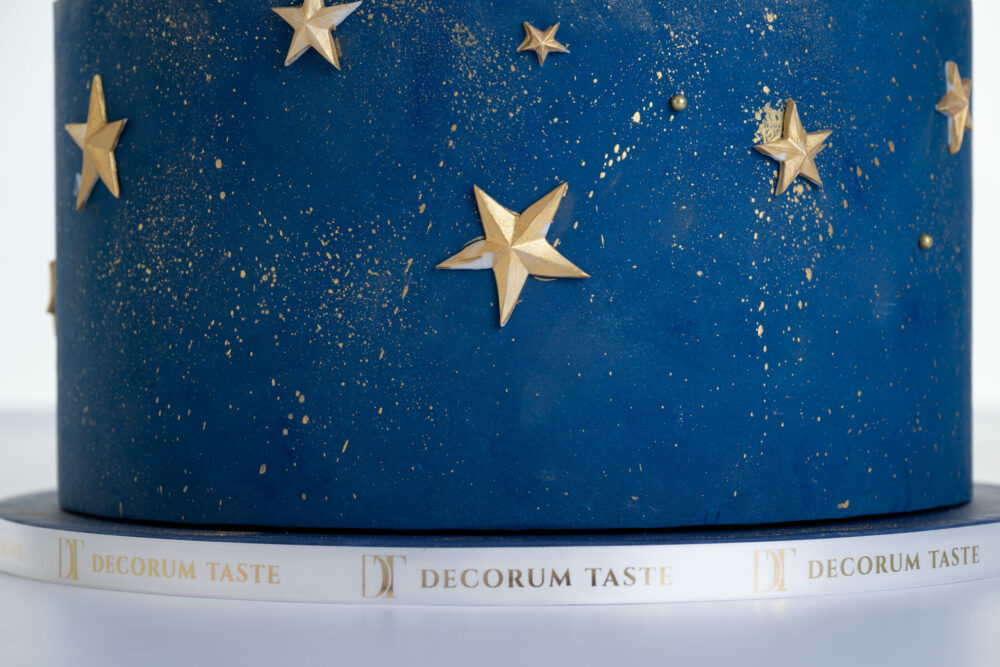 Bottom tier of luxury contemporary, customisable, celebration cake, Cosmos Star by Decorum Taste, in deep blue with flecks of edible gold and golden stars. The cake board is wrapped in a Decorum Taste logo ribbon.