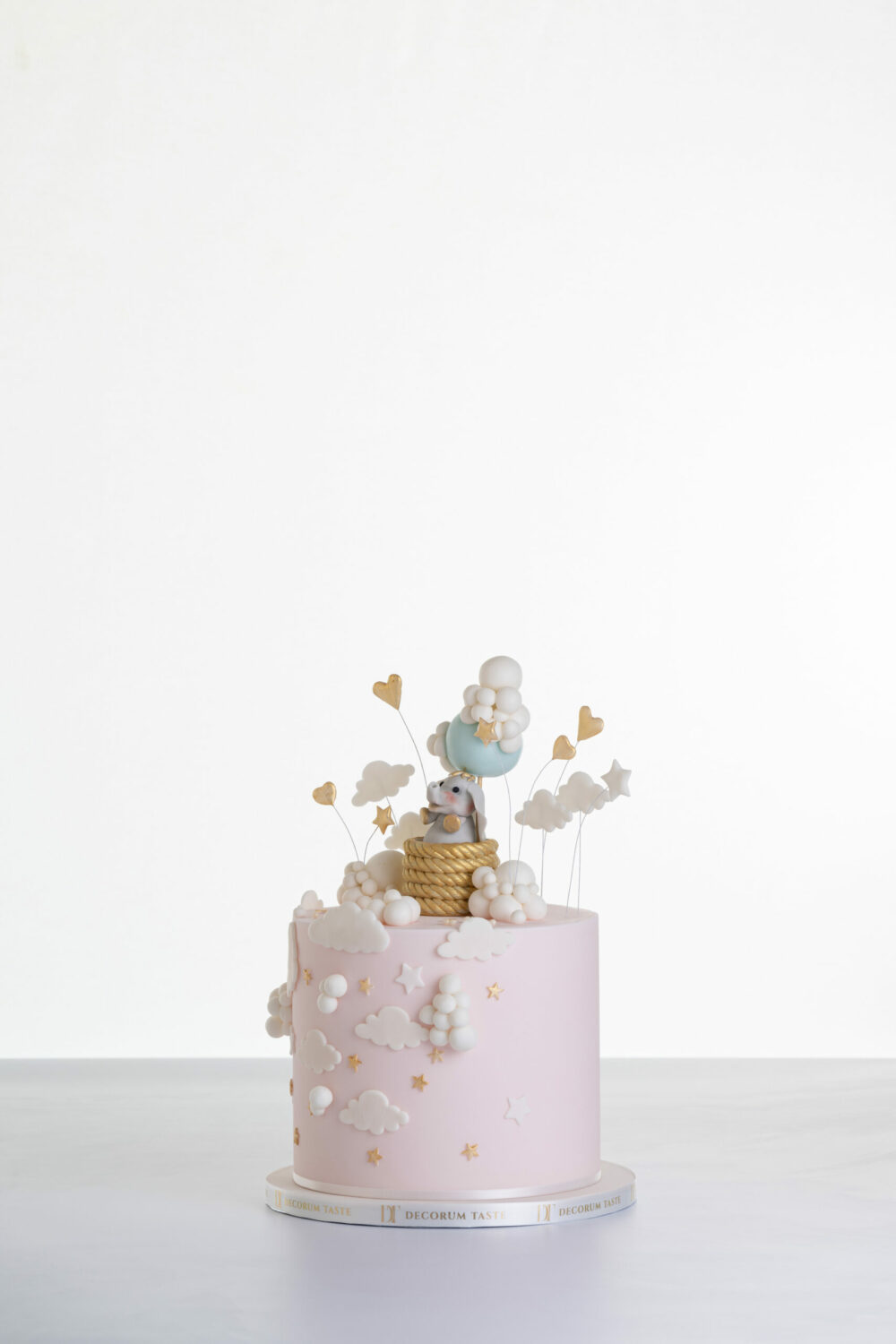 Single tier luxury contemporary, customisable, celebration cake, Cute Elephant by Decorum Taste, in blush pink with hand-crafted elephant in sugar paste sitting on top balancing a balloon, with embellishments of clouds, stars and hearts.