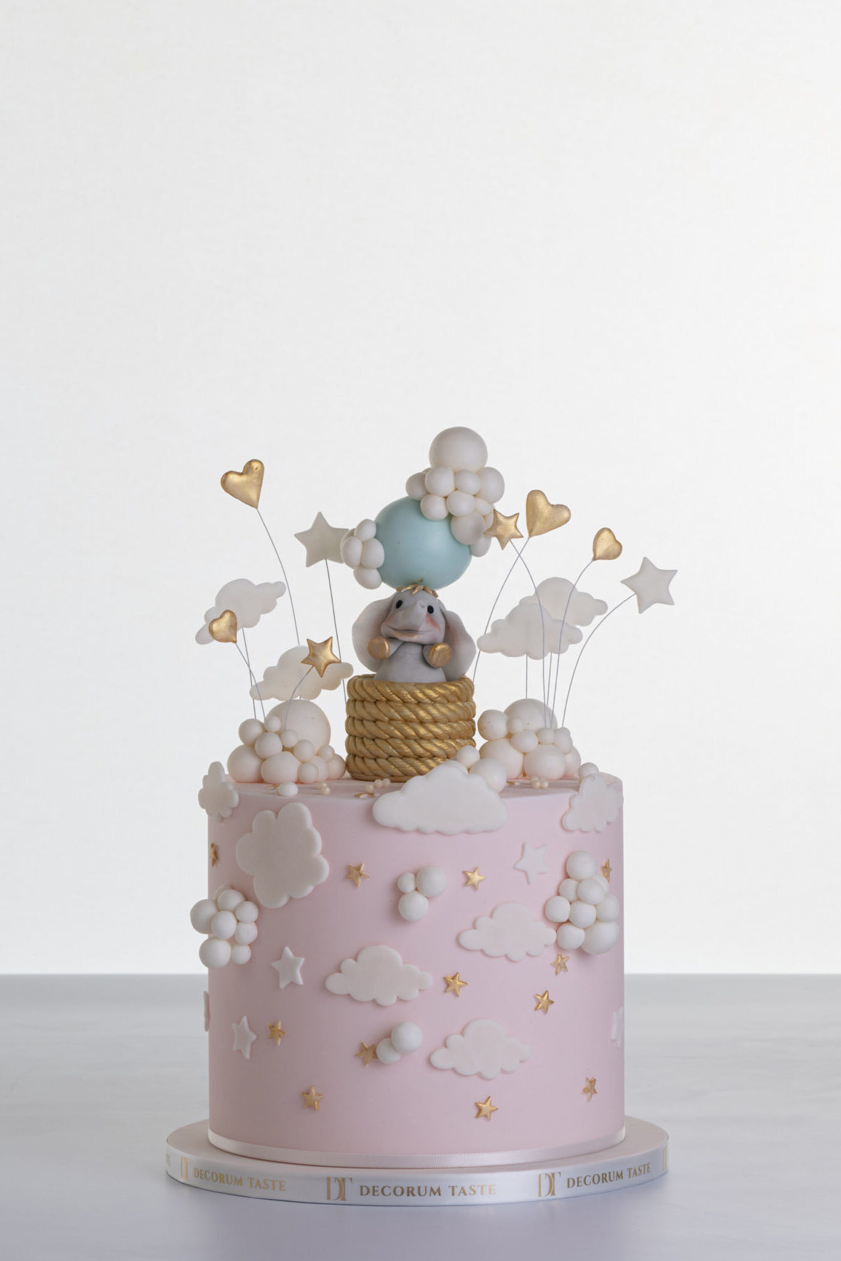 Single tier luxury contemporary, customisable, celebration cake, Cute Elephant by Decorum Taste, in blush pink with hand-crafted elephant in sugar paste sitting on top balancing a balloon, with embellishments of clouds, stars and hearts.