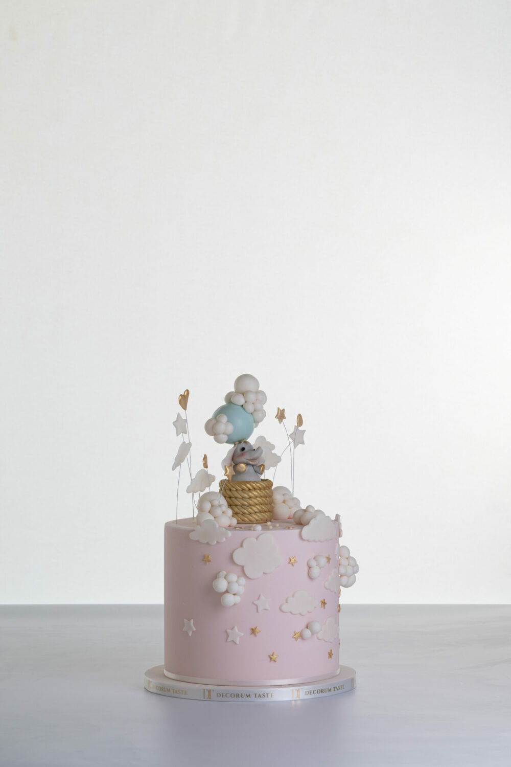 Single tier luxury contemporary, customisable, celebration cake, Cute Elephant by Decorum Taste, in blush pink with hand-crafted elephant in sugar paste sitting on top balancing a balloon, with embellishments of clouds, stars and hearts.