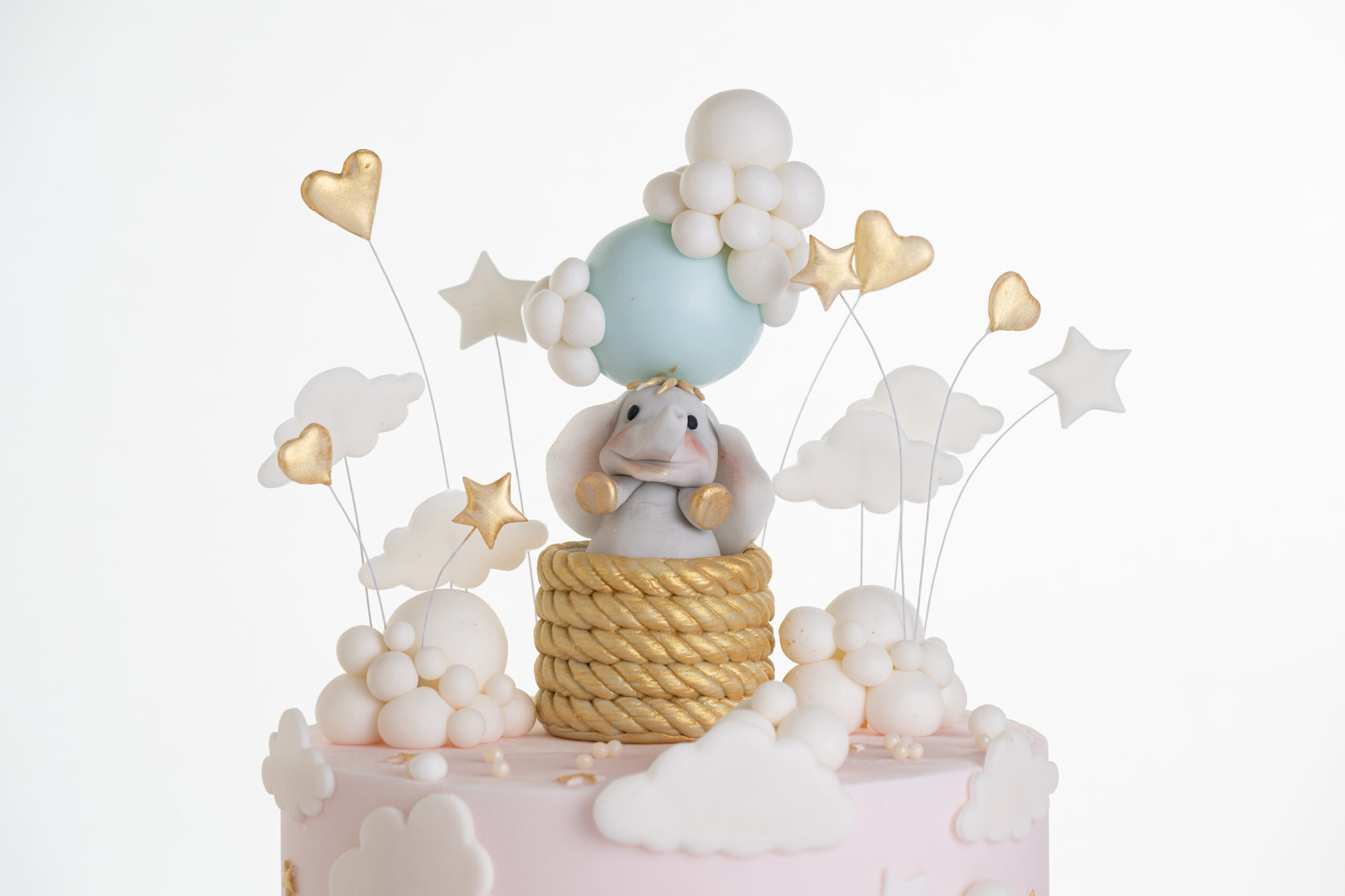 Top of luxury contemporary, customisable, celebration cake, Cute Elephant by Decorum Taste, in blush pink with hand-crafted elephant in sugar paste sitting on top balancing a balloon, with embellishments of clouds, stars and hearts.