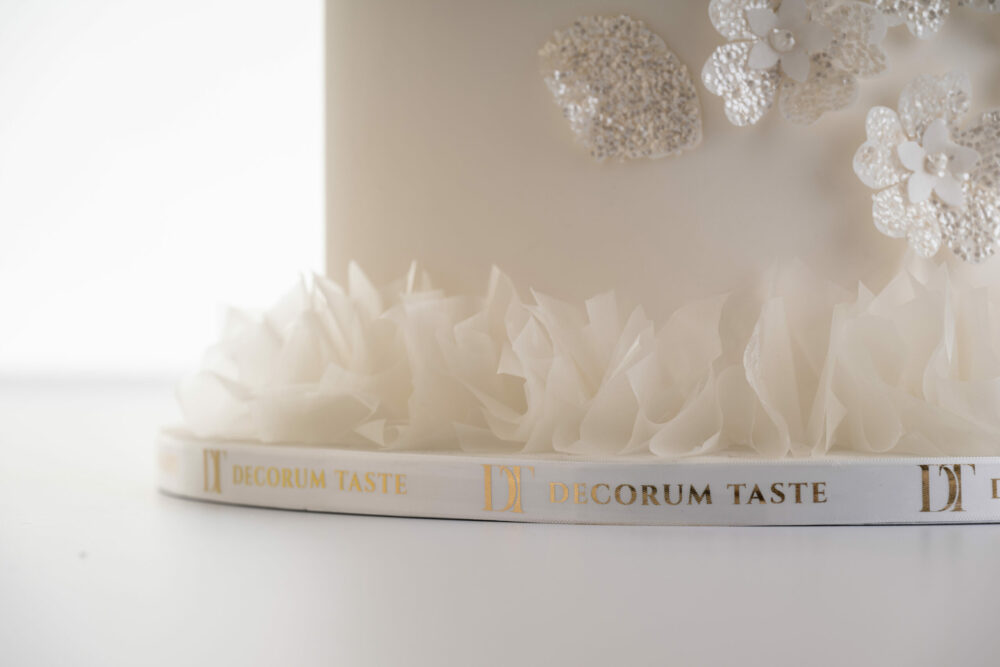 Base of luxury customisable wedding cake, Daphne, with rice paper frill, glittery floral decoration and a branded ribbon with the DT logo and words Decorum Taste in repeat around the cake board.