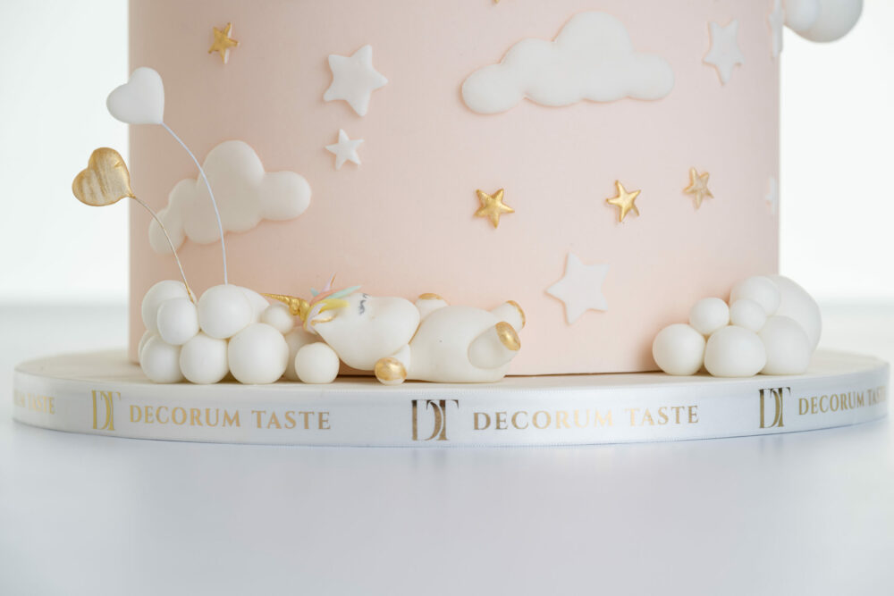 Bottom tier of luxury contemporary, customisable celebration cake, Floating Unicorn by Decorum Taste, decorated in blush pink with embellishments of white clouds, stars and a unicorn asleep at the base. The cake board is wrapped in a ribbon with the Decorum Taste logo.