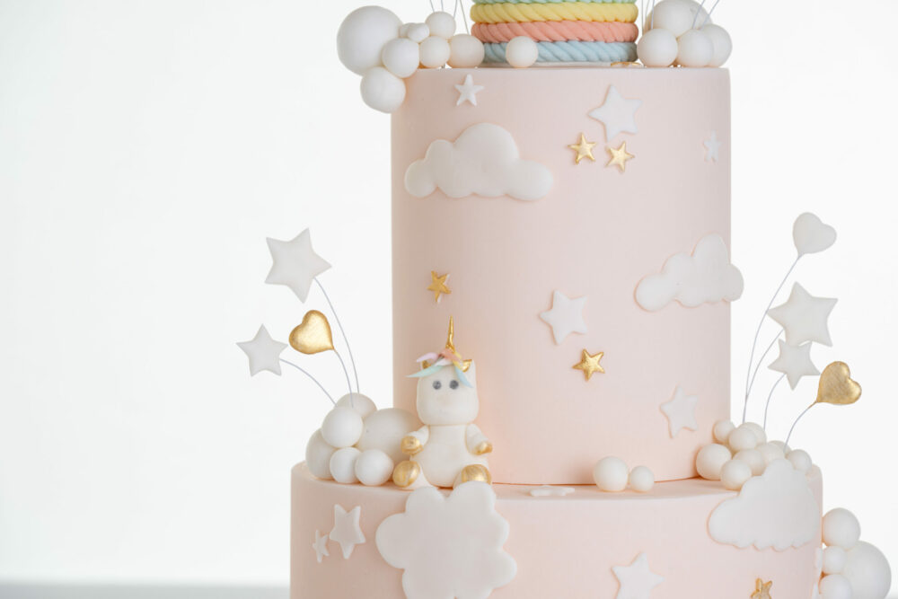 Detail of luxury contemporary, customisable celebration cake, Floating Unicorn by Decorum Taste, decorated in blush pink with embellishments of white clouds, stars with a unicorn at the top sitting in a pastel coloured hot air balloon and another resting on the top of the first tier.