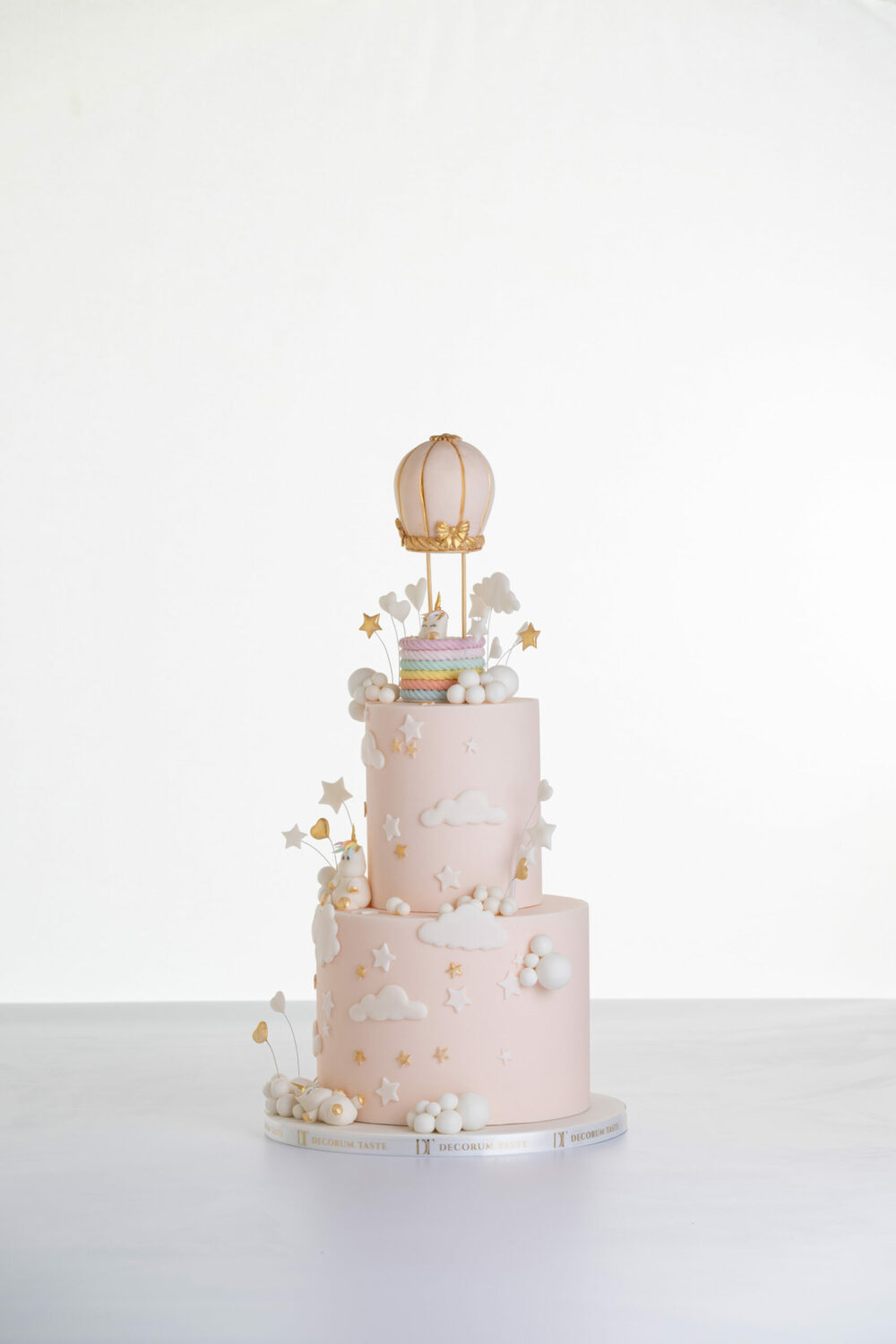 Two tier luxury contemporary, customisable celebration cake, Floating Unicorn by Decorum Taste, decorated in blush pink with embellishments of white clouds, stars with a unicorn at the top sitting in a pastel coloured hot air balloon and another resting on the top of the first tier and a third asleep at the bottom.