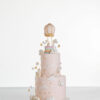 Two tier luxury contemporary, customisable celebration cake, Floating Unicorn by Decorum Taste, decorated in blush pink with embellishments of white clouds, stars with a unicorn at the top sitting in a pastel coloured hot air balloon and another resting on the top of the first tier and a third asleep at the bottom.