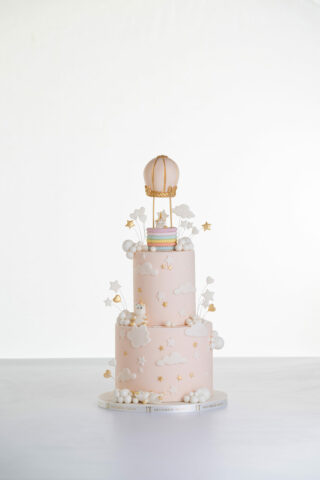 Two tier luxury contemporary, customisable celebration cake, Floating Unicorn by Decorum Taste, decorated in blush pink with embellishments of white clouds, stars with a unicorn at the top sitting in a pastel coloured hot air balloon and another resting on the top of the first tier and a third asleep at the bottom.