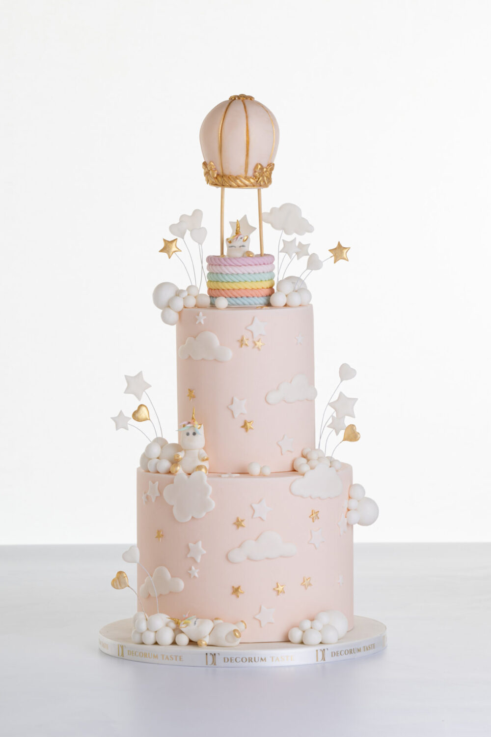 Two tier luxury contemporary, customisable celebration cake, Floating Unicorn by Decorum Taste, decorated in blush pink with embellishments of white clouds, stars with a unicorn at the top sitting in a pastel coloured hot air balloon and another resting on the top of the first tier and a third asleep at the bottom.