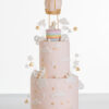 Two tier luxury contemporary, customisable celebration cake, Floating Unicorn by Decorum Taste, decorated in blush pink with embellishments of white clouds, stars with a unicorn at the top sitting in a pastel coloured hot air balloon and another resting on the top of the first tier and a third asleep at the bottom.