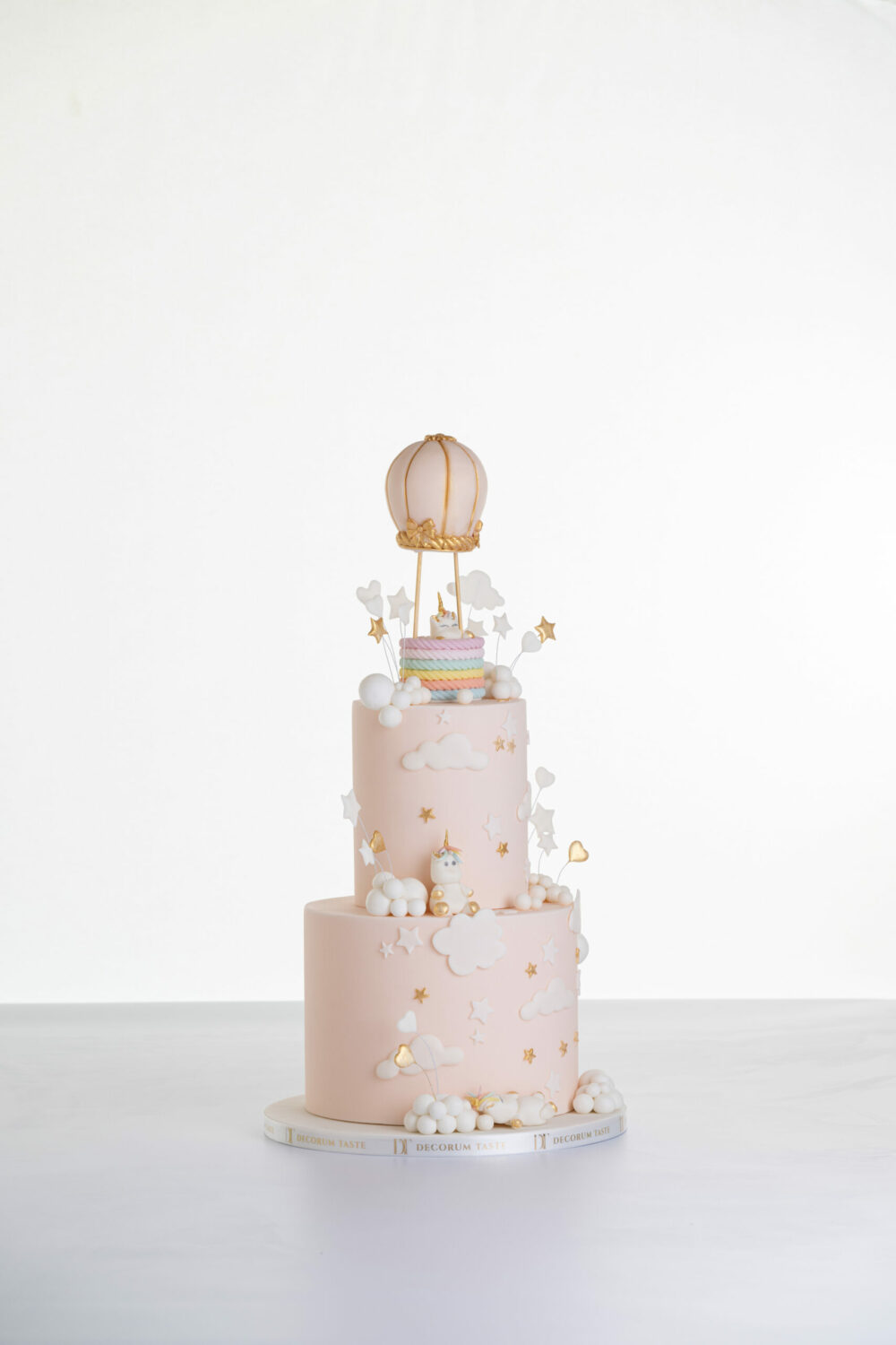 Two tier luxury contemporary, customisable celebration cake, Floating Unicorn by Decorum Taste, decorated in blush pink with embellishments of white clouds, stars with a unicorn at the top sitting in a pastel coloured hot air balloon and another resting on the top of the first tier and a third asleep at the bottom.