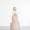 Two tier luxury contemporary, customisable celebration cake, Floating Unicorn by Decorum Taste, decorated in blush pink with embellishments of white clouds, stars with a unicorn at the top sitting in a pastel coloured hot air balloon and another resting on the top of the first tier and a third asleep at the bottom.