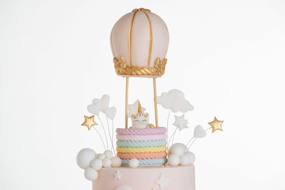 Top tier of luxury contemporary, customisable celebration cake, Floating Unicorn by Decorum Taste, decorated in blush pink with embellishments of white clouds, stars and a unicorn at the top sitting in a pastel coloured hot air balloon.