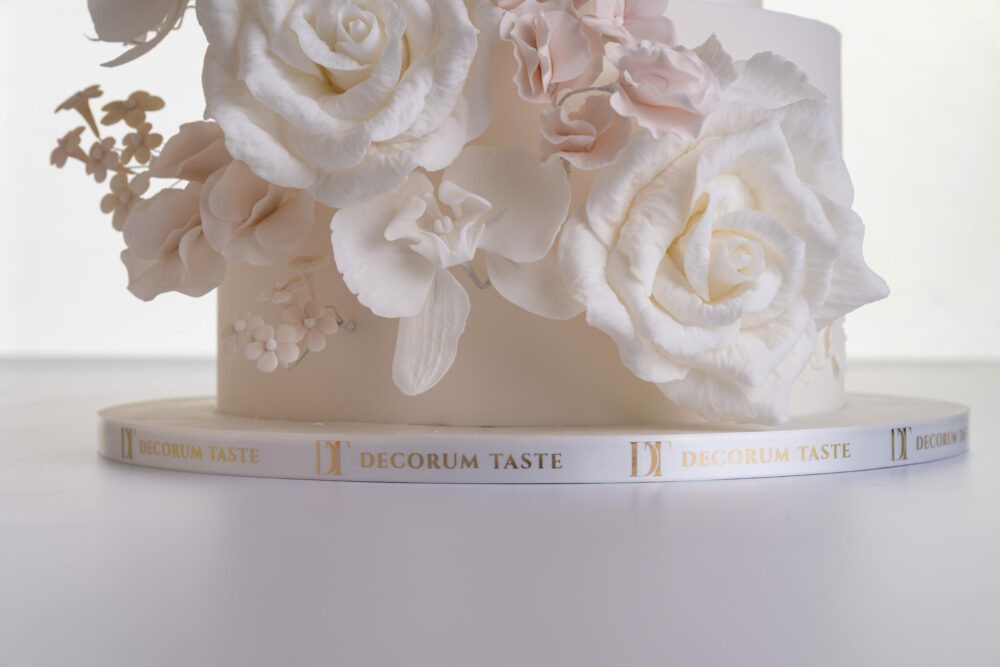 Bottom tier of luxury contemporary, customisable, wedding cake, Floral Cascade by Decorum Taste, decorated with hand-crafted sugar roses, sweet peas and other petals in ivory and blush pink tones. A branded decorative ribbon wraps the side of the cake board with the Decorum Taste logo.