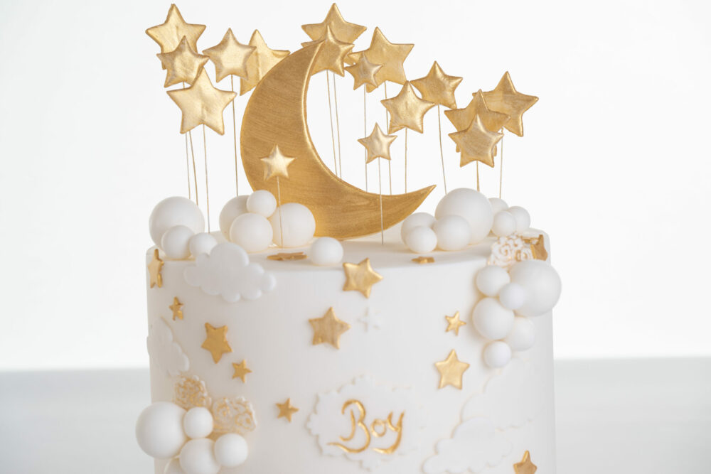 Top of luxury contemporary, customisable, celebration cake, Moon Baby by Decorum Taste, in ivory with large golden moon, stars and clouds.