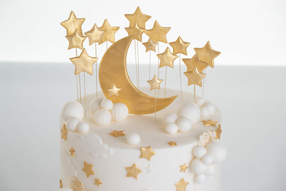 Top of luxury contemporary, customisable, celebration cake, Moon Baby by Decorum Taste, in ivory with large golden moon, stars and clouds.