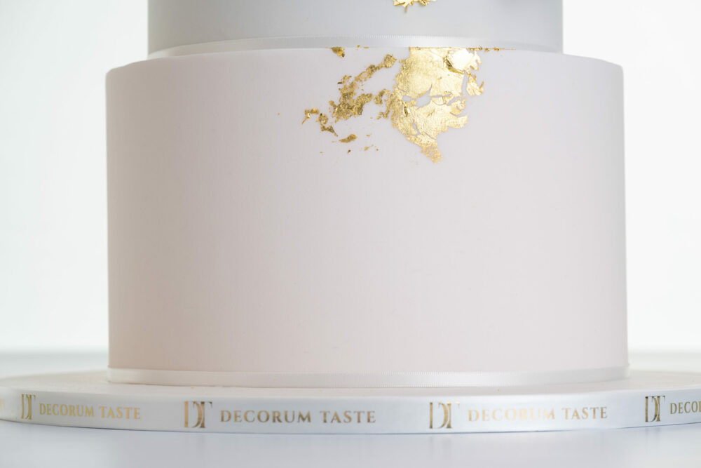 Bottom tier of luxury contemporary, customisable, wedding cake, Orchid Heaven by Decorum Taste, in blush pink and brushes of edible gold leaf. The side of the cake board is decorated in a Decorum Tase logo branded ribbon.