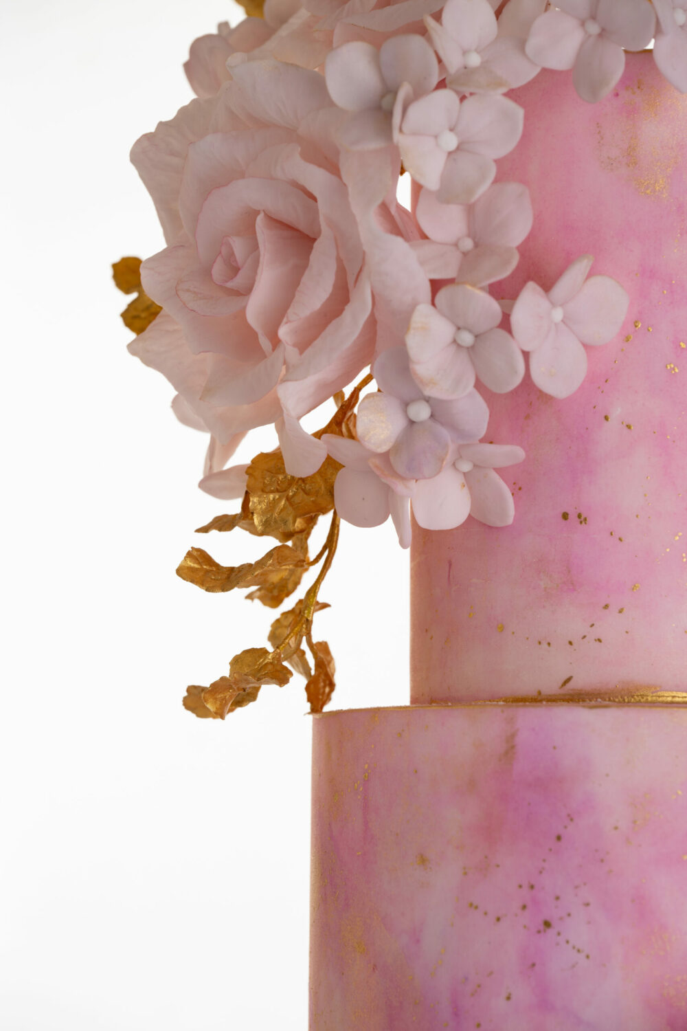 Side detail of luxury customisable contemporary wedding cake, Painted Blush by Decorum Taste, in hand-painted pink tones with gold flecks and a bouquet of hand-crafted sugar roses, hydrangea petals and trailing gold leaves.