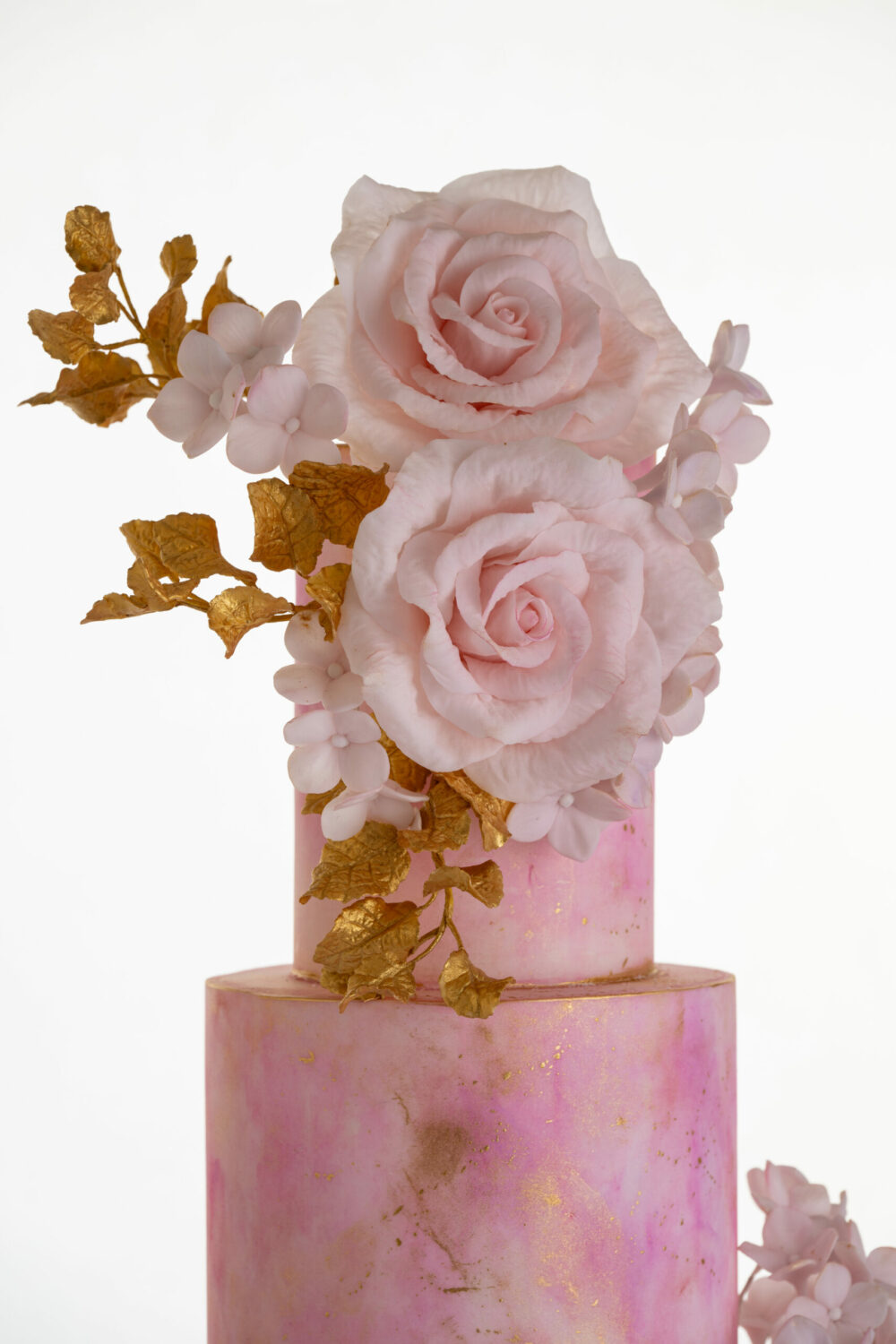 Top two tiers of luxury customisable contemporary wedding cake, Painted Blush, by Decorum Taste in pink hand-painted tones with a bouquet of large hand-crafted sugar roses, hydrangea petals and golden leaves.