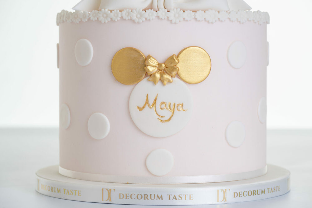 Bottom tier of luxury contemporary, customisable, celebration cake, Polka Mouse by Decorum Taste, with decorative edible circle embellishments, a pretty floral trim and cartoon mouse shape made from circles with child's name in the middle and gold bow. The cake board is wrapped with a Decorum Taste logo branded ribbon.