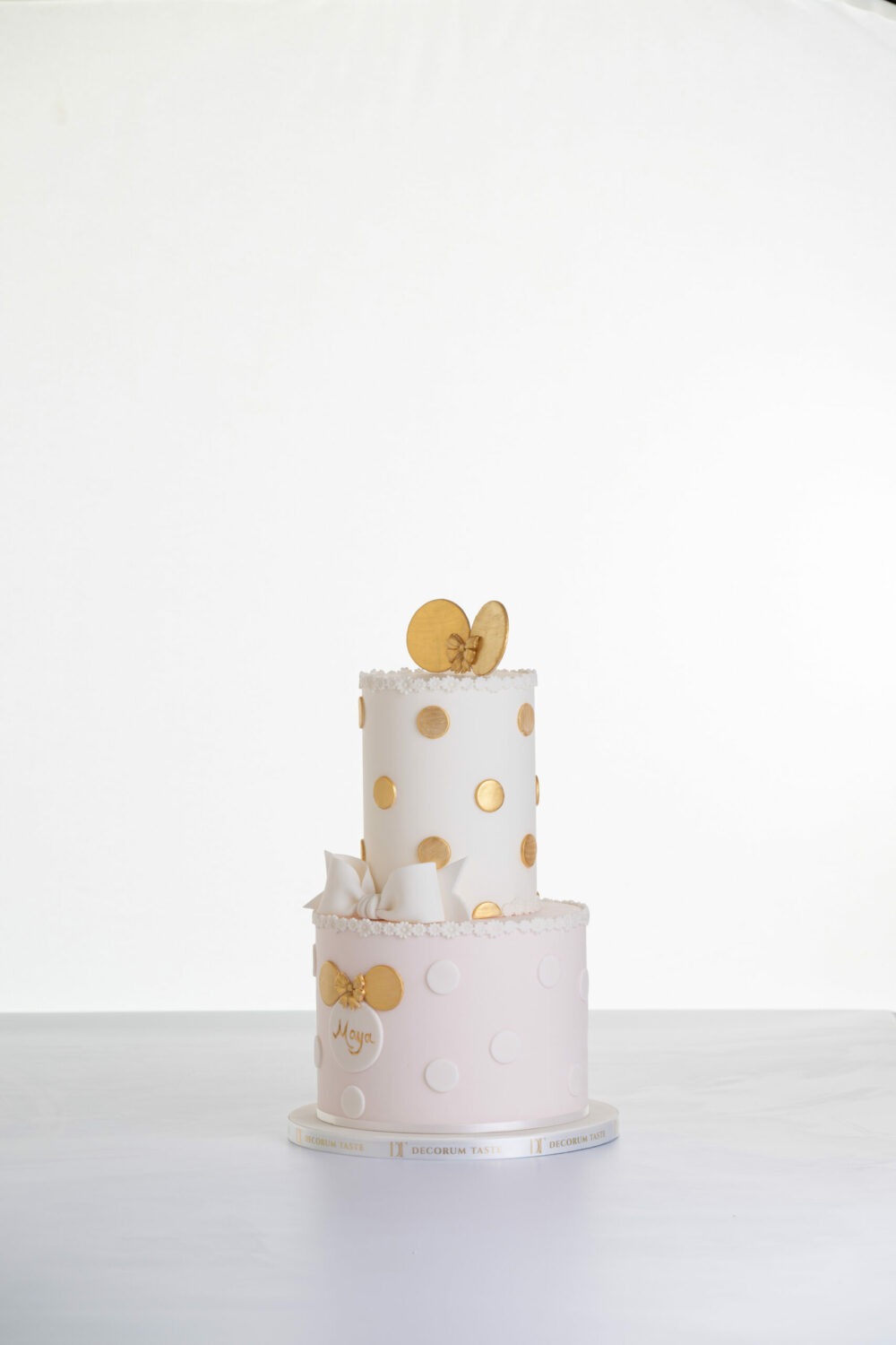 Two tier luxury contemporary, customisable, celebration cake, Polka Mouse by Decorum Taste, with decorative edible gold leaf circle embellishments, a pretty floral trim, cartoon mouse ears and sugar paste bow.