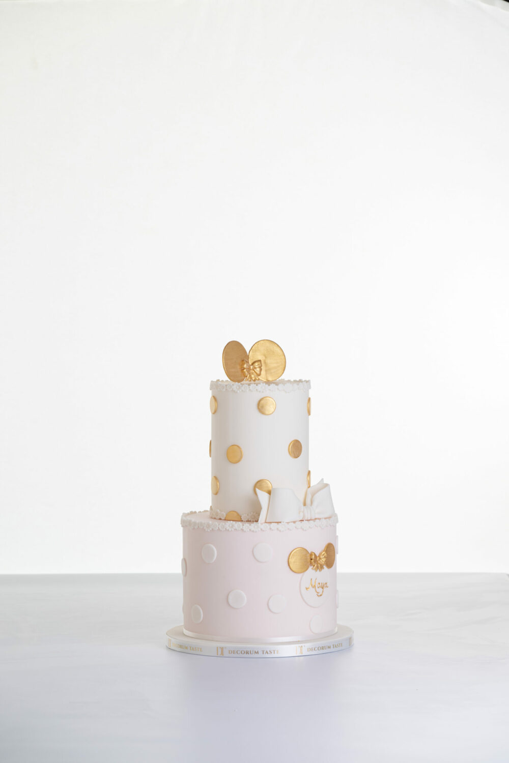 Two tier luxury contemporary, customisable, celebration cake, Polka Mouse by Decorum Taste, with decorative edible gold leaf circle embellishments, a pretty floral trim, cartoon mouse ears and sugar paste bow.