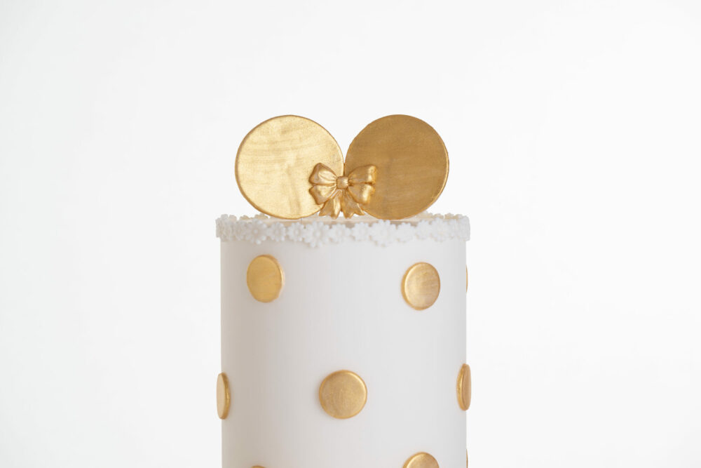 Top tier of luxury contemporary, customisable, celebration cake, Polka Mouse by Decorum Taste, with decorative edible gold leaf circle embellishments, a pretty floral trim and round mouse ears on top in gold with bow.