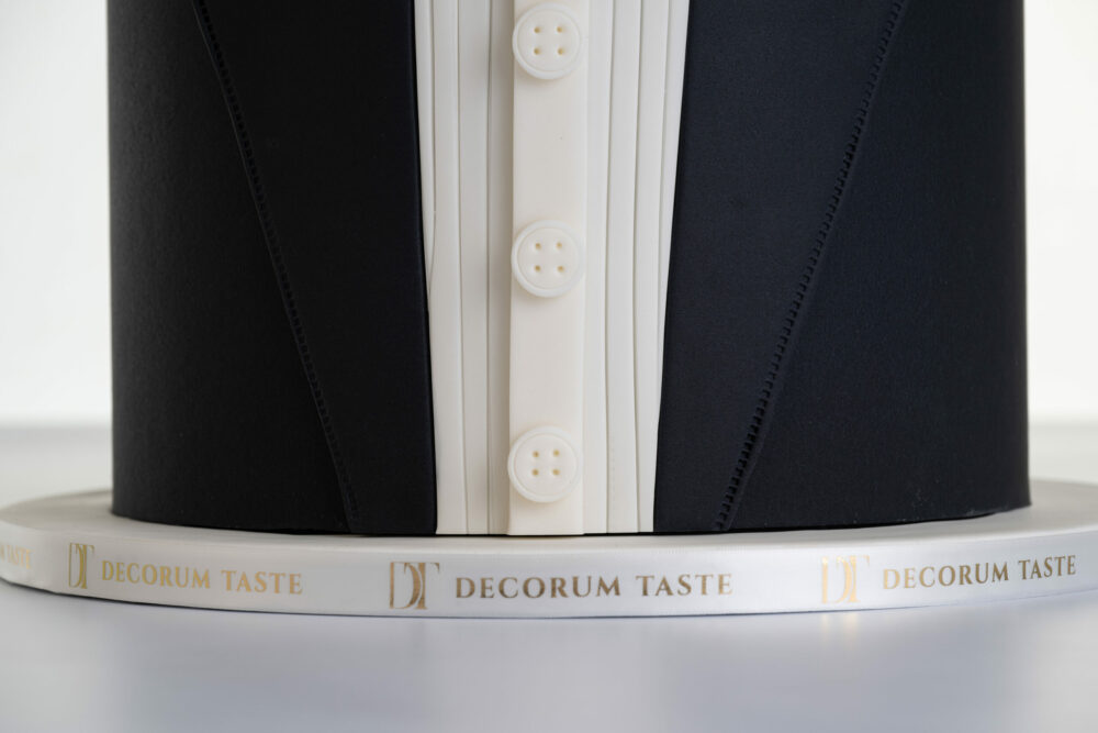 Bottom of luxury contemporary, customisable, celebration cake, Tux Gent by Decorum Taste, decorated in black the white of a formal shirt, tuxedo jacket and bow tie. The cake base is wrapped in a ribbon with the Decorum Taste logo.