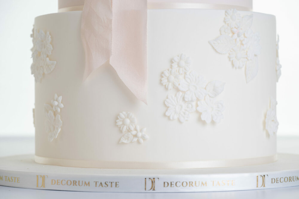 Detail of bottom tier of luxury contemporary, customisable, wedding cake, Vintage Beauty by Decorum Taste with decorative floral embellishments and the tail of a pink silk ribbon. The side of the cake board is wrapped in a Decorum Taste logo branded ribbon.