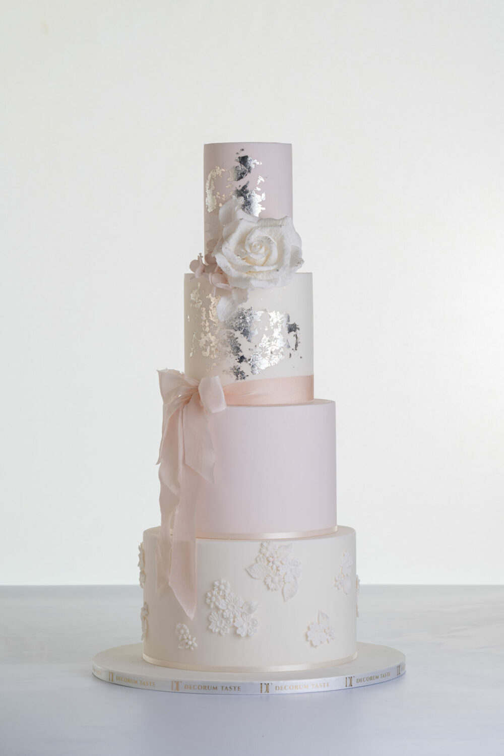 Four tier luxury contemporary, customisable, wedding cake, Vintage Beauty by Decorum Taste with large hand-crafted sugar rose in ivory, brushed edible silver leaf, decorative floral embellishments and a pink silk bow.