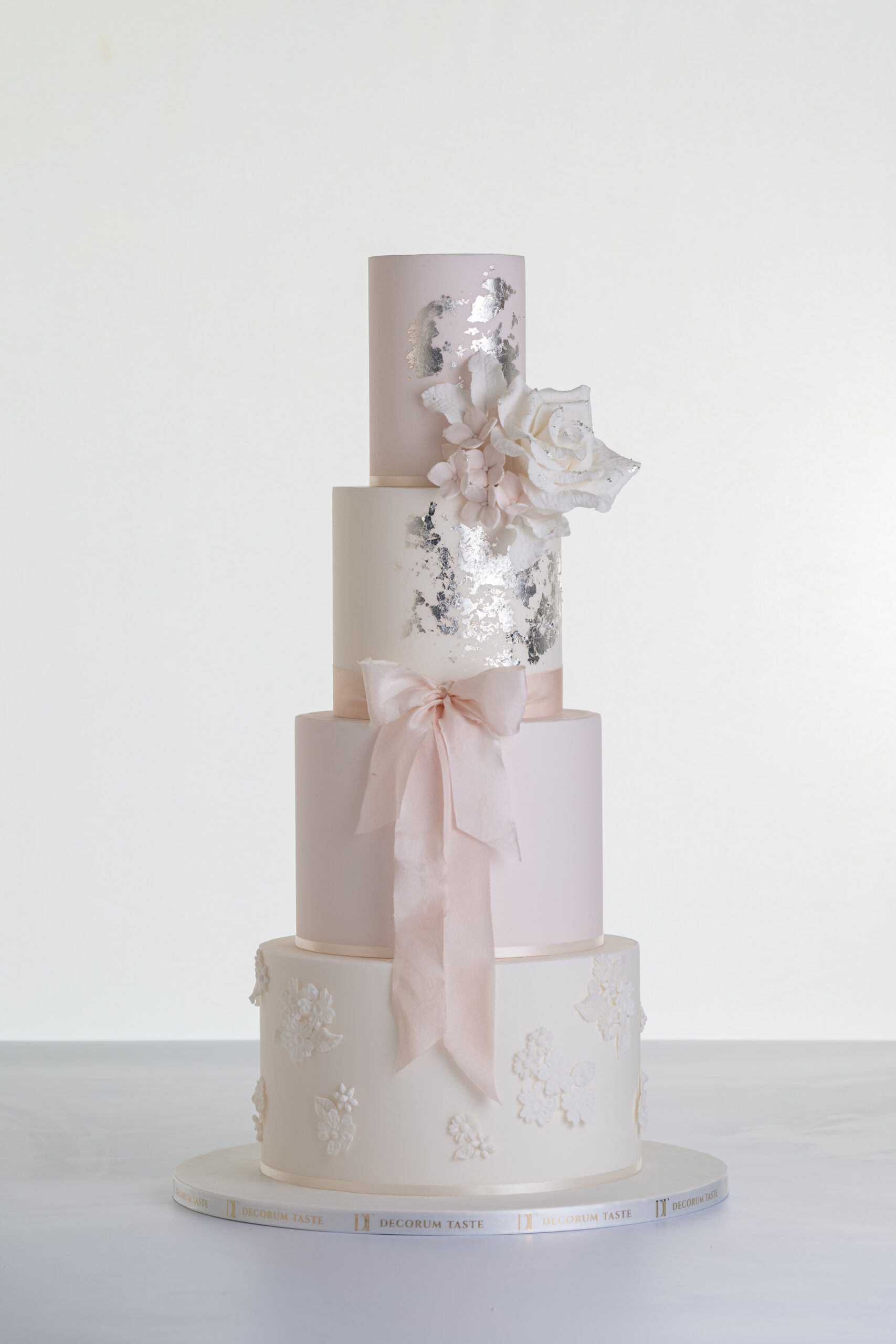 Four tier luxury contemporary, customisable, wedding cake, Vintage Beauty by Decorum Taste with large hand-crafted sugar rose in ivory, brushed edible silver leaf, decorative floral embellishments and a pink silk bow.