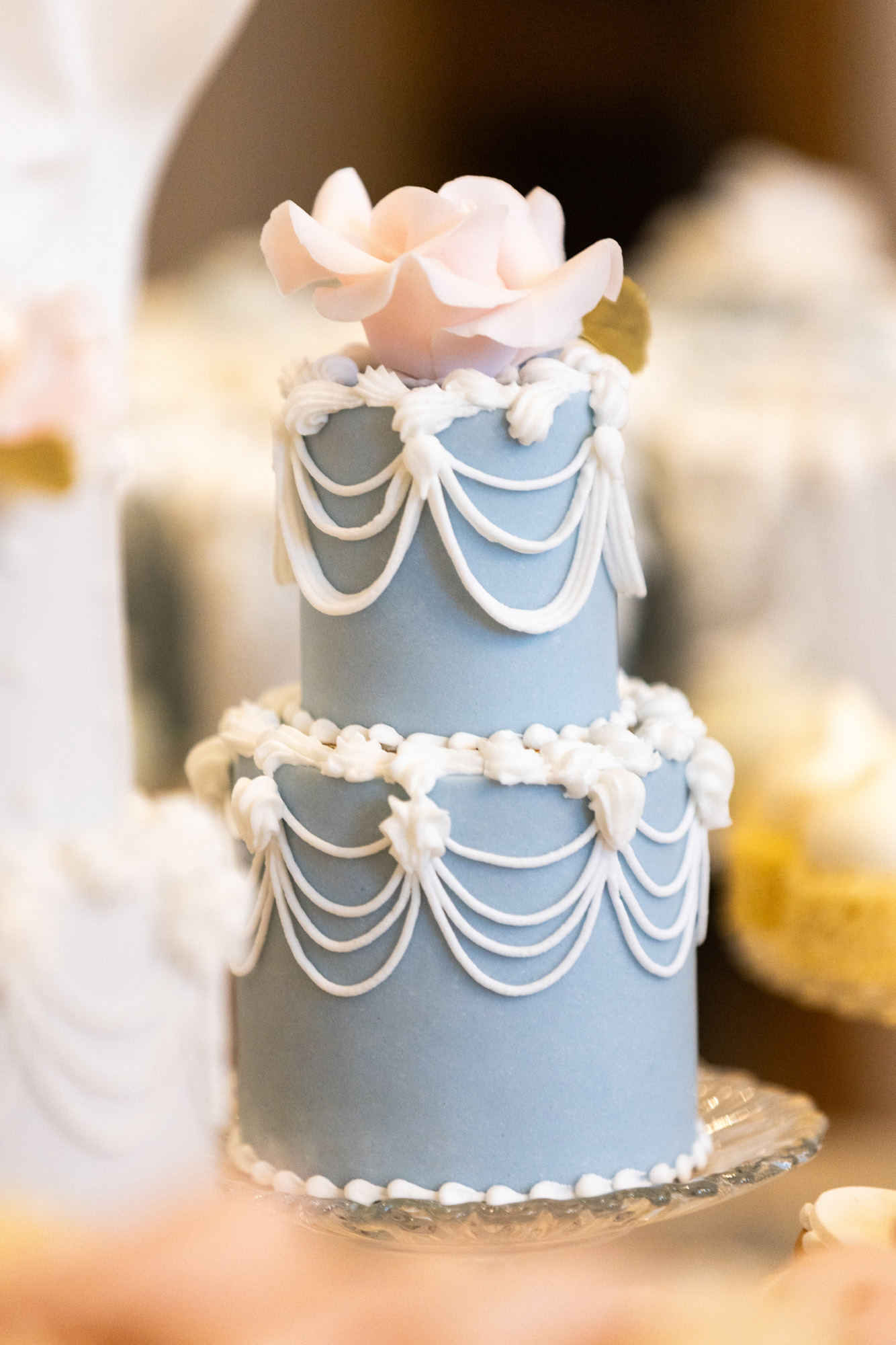 By Yevnig luxury bespoke two-tier petite favour cake in blue Royal icing with piped swags and details with a hand-crafted rose in blush pink on top.