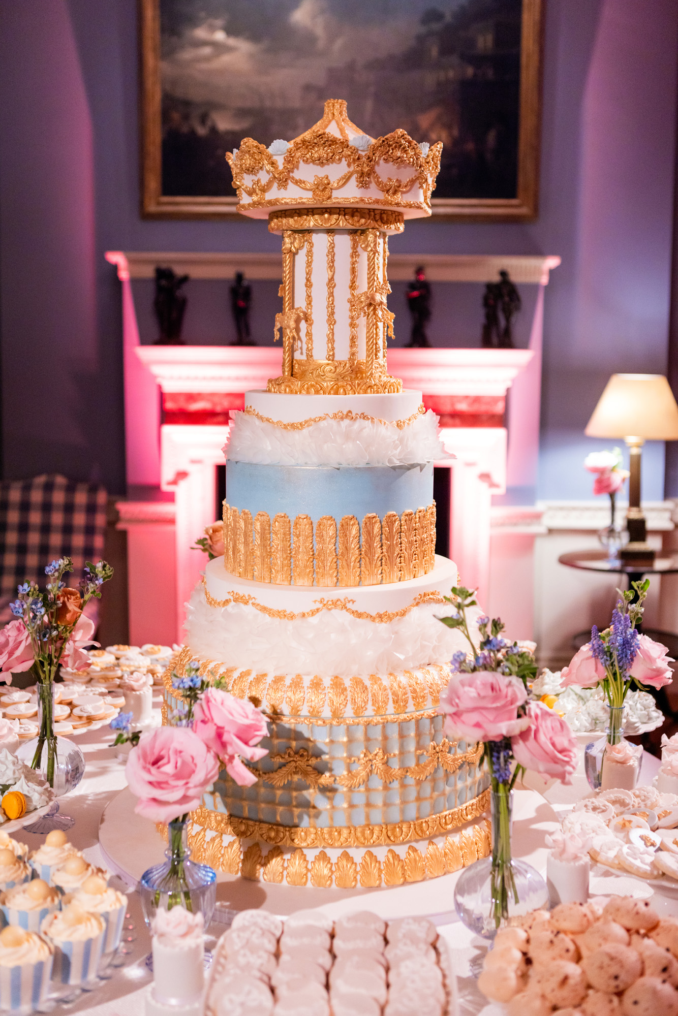 Luxury bespoke dessert table for child birthday, Christening, baby shower, baby naming ceremony, The Carousel, By Yevnig with statement centrepiece cake in pale blue, blush pink and gold detailing replicating a vintage fairground carousel. The table is decorated with petite cakes, macarons, luxury biscuits and other sweet treats